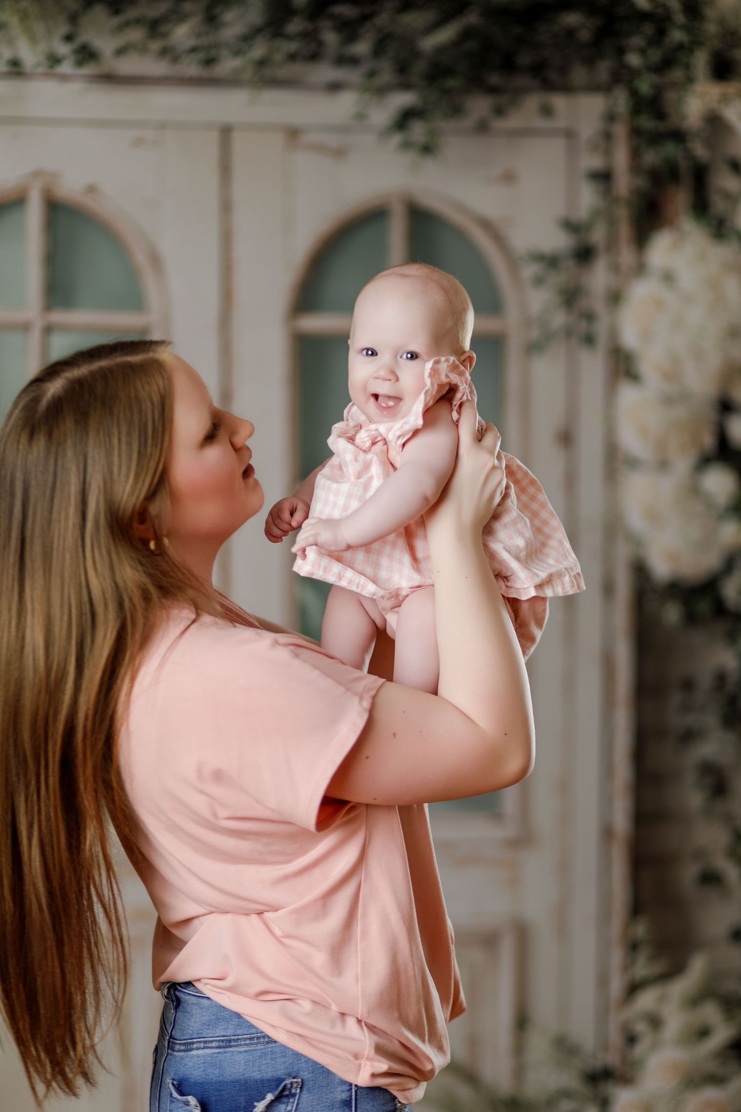 Mother in pink shirt holding baby girl up near floral decorated door backdrop.
