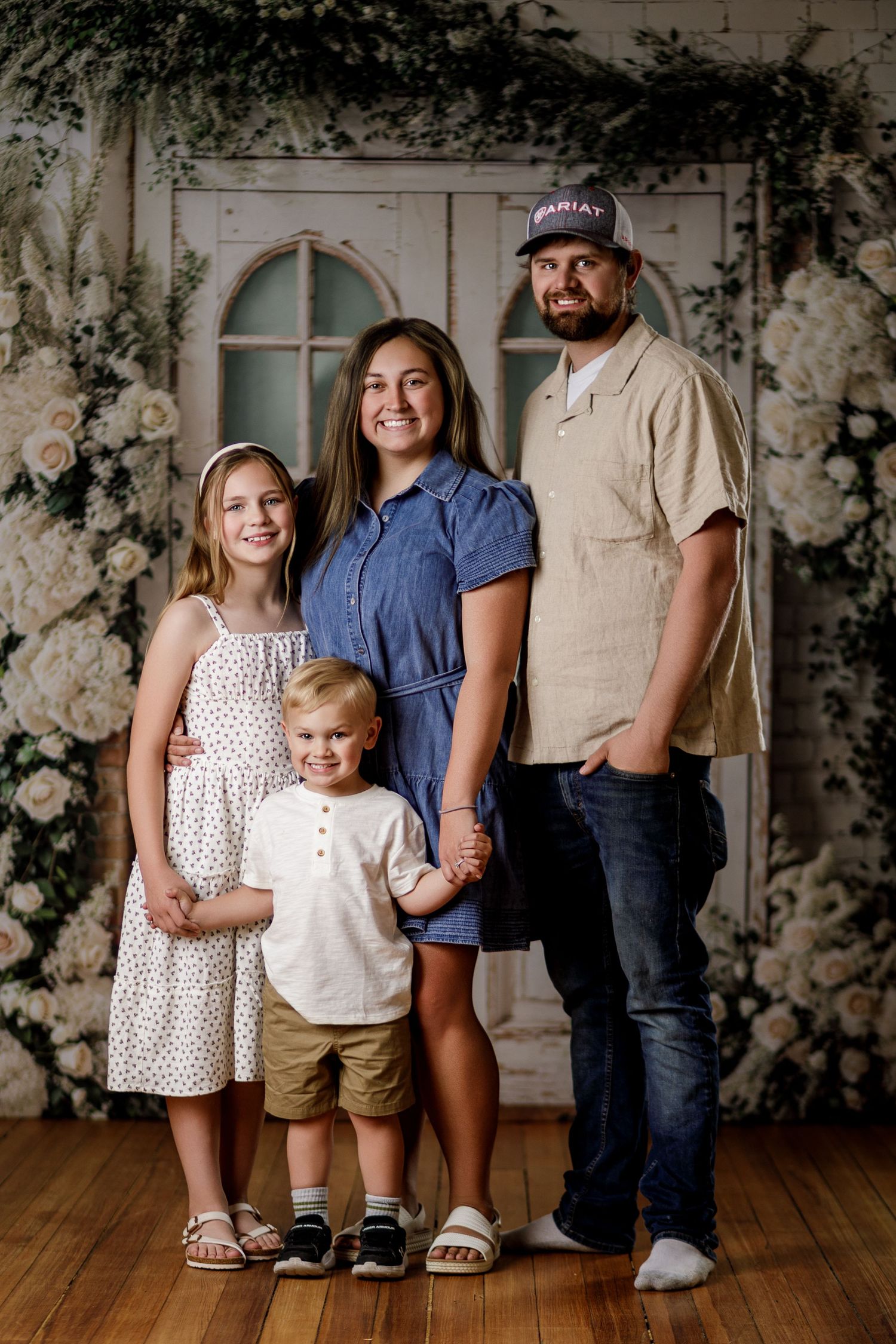 Family of four posing for a portrait in front of a floral backdrop with white roses and vintage door decor.