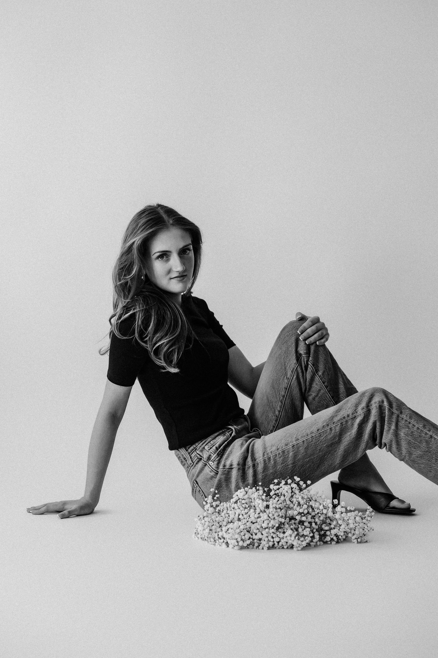 Black and white photo of young woman sitting on floor in black top and jeans with baby's breath flowers, leaning back casually.