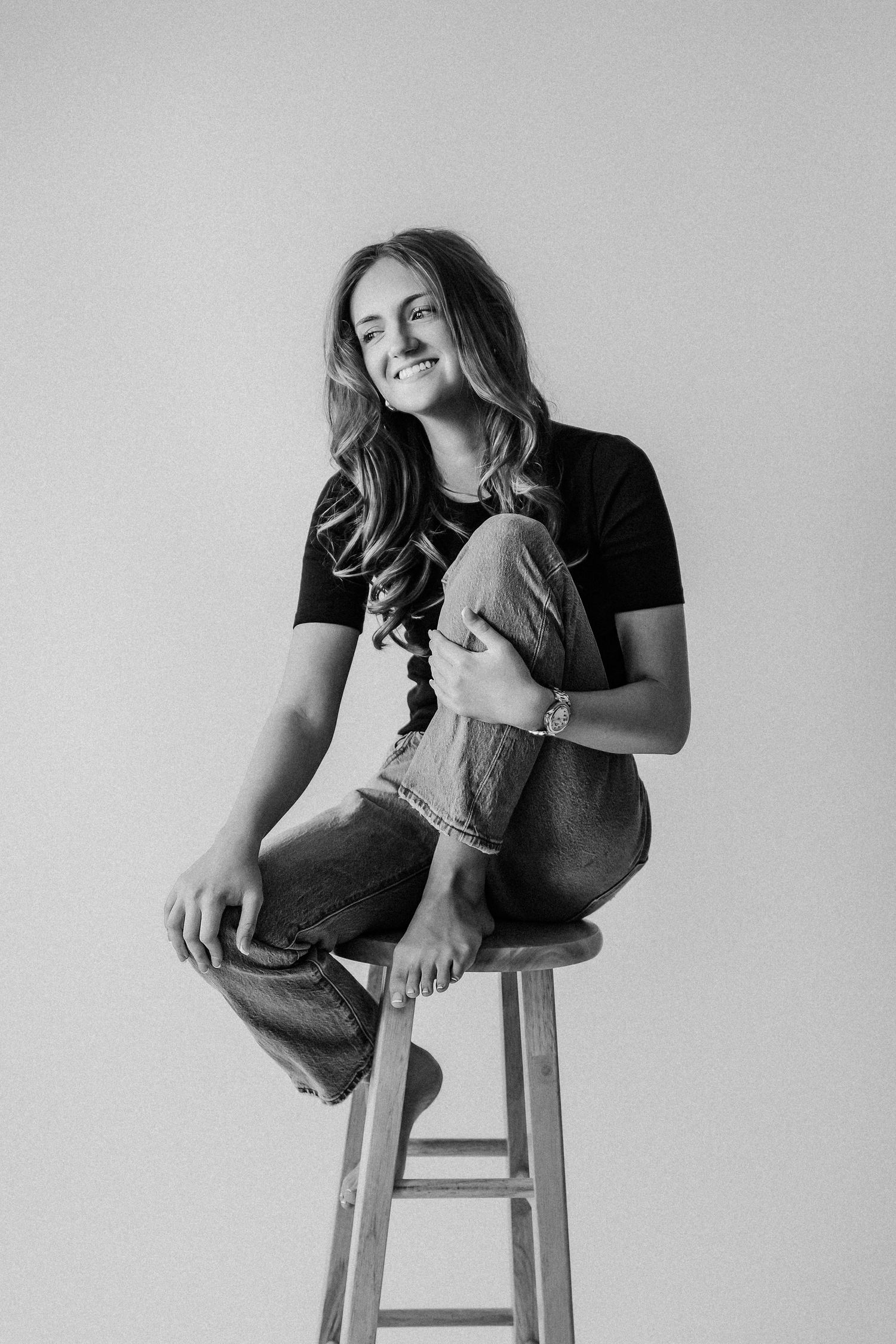 Young woman smiling on wooden stool, black tee and jeans, black and white portrait photo.