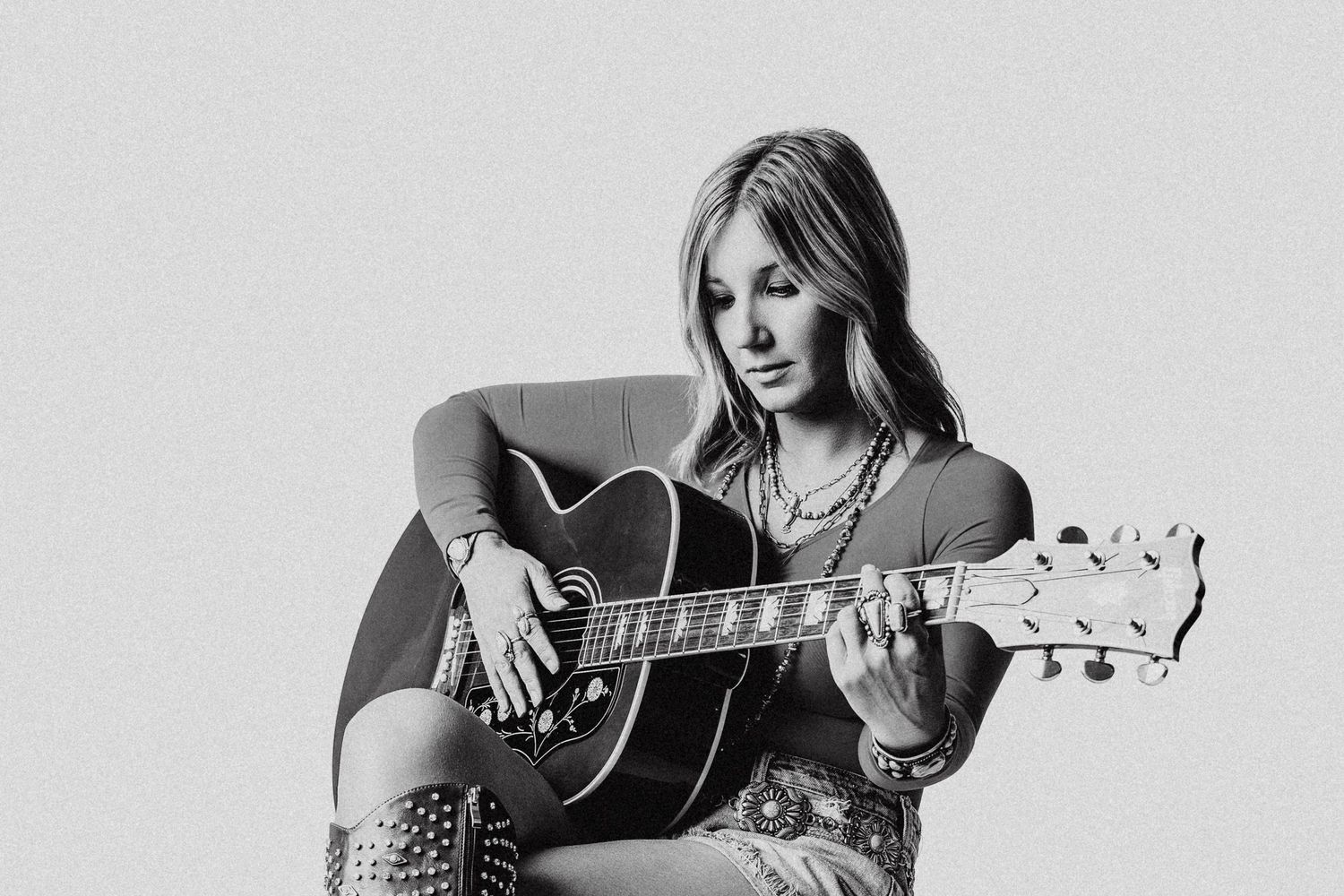 Black and white photo of a young woman playing acoustic guitar while seated, wearing bohemian style clothing and layered necklaces.