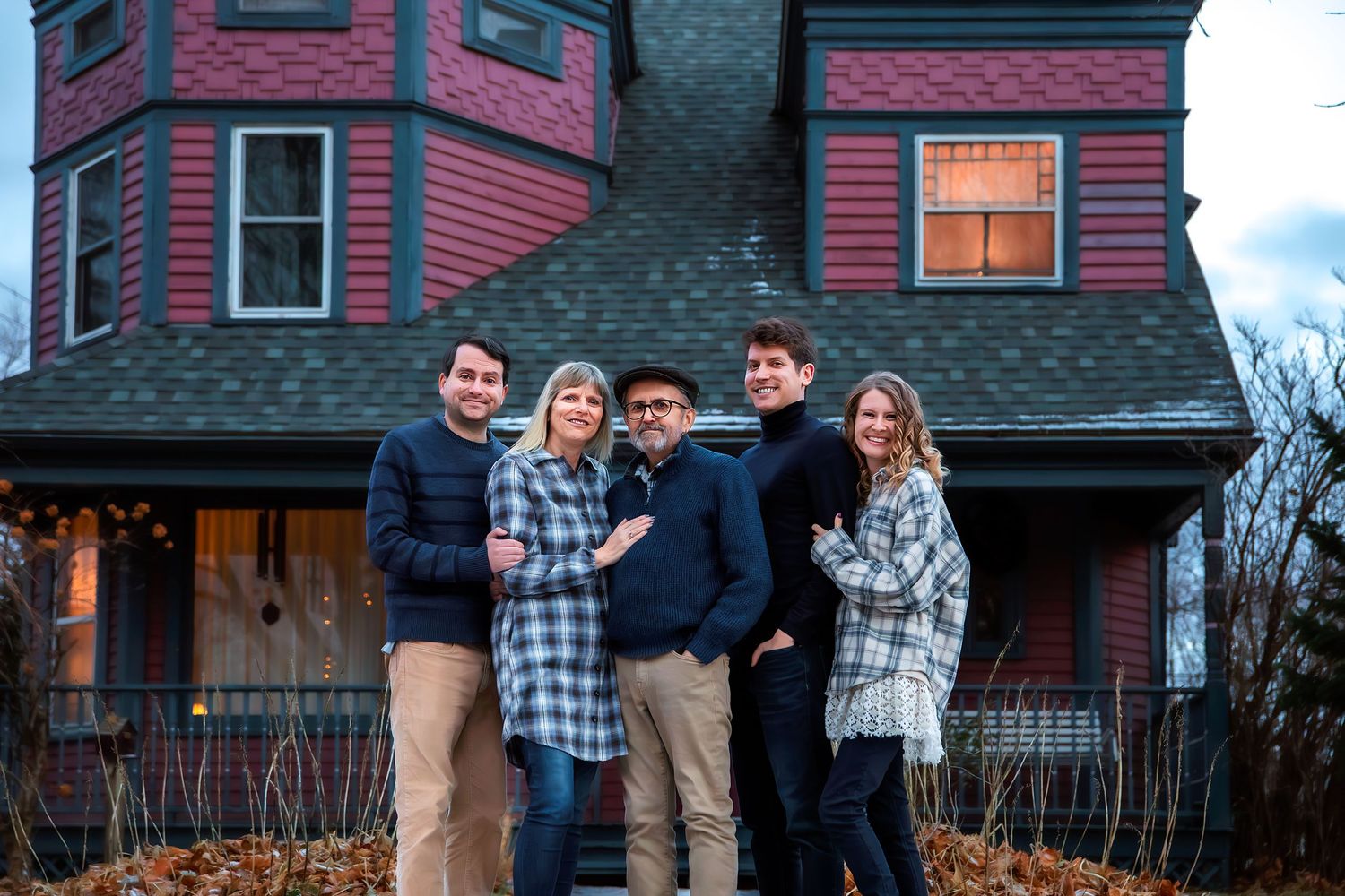 A group of people pose together in front of a red house during sunset on a winter evening.