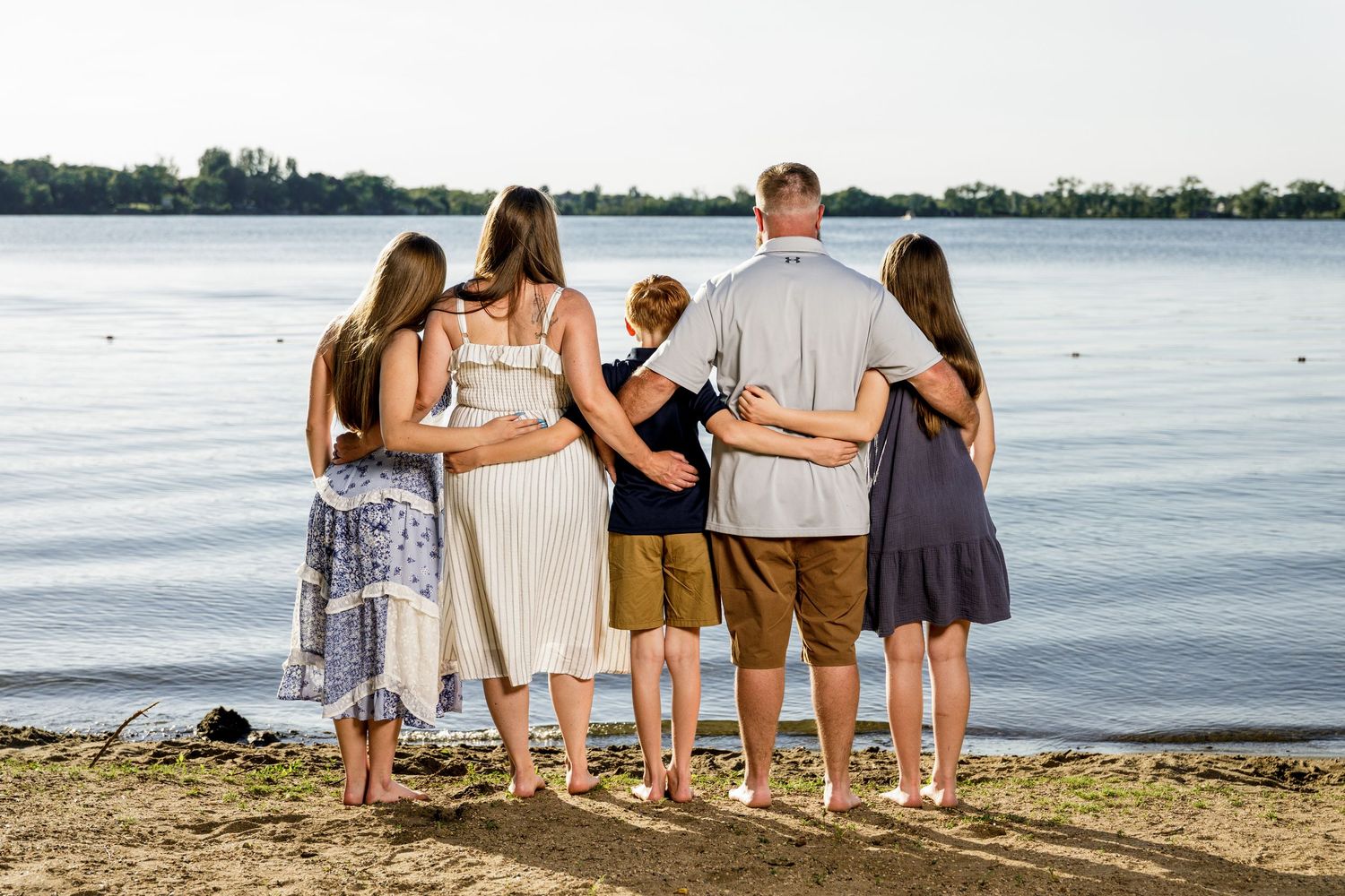 Family of six standing together from behind on sandy beach shore overlooking calm lake.