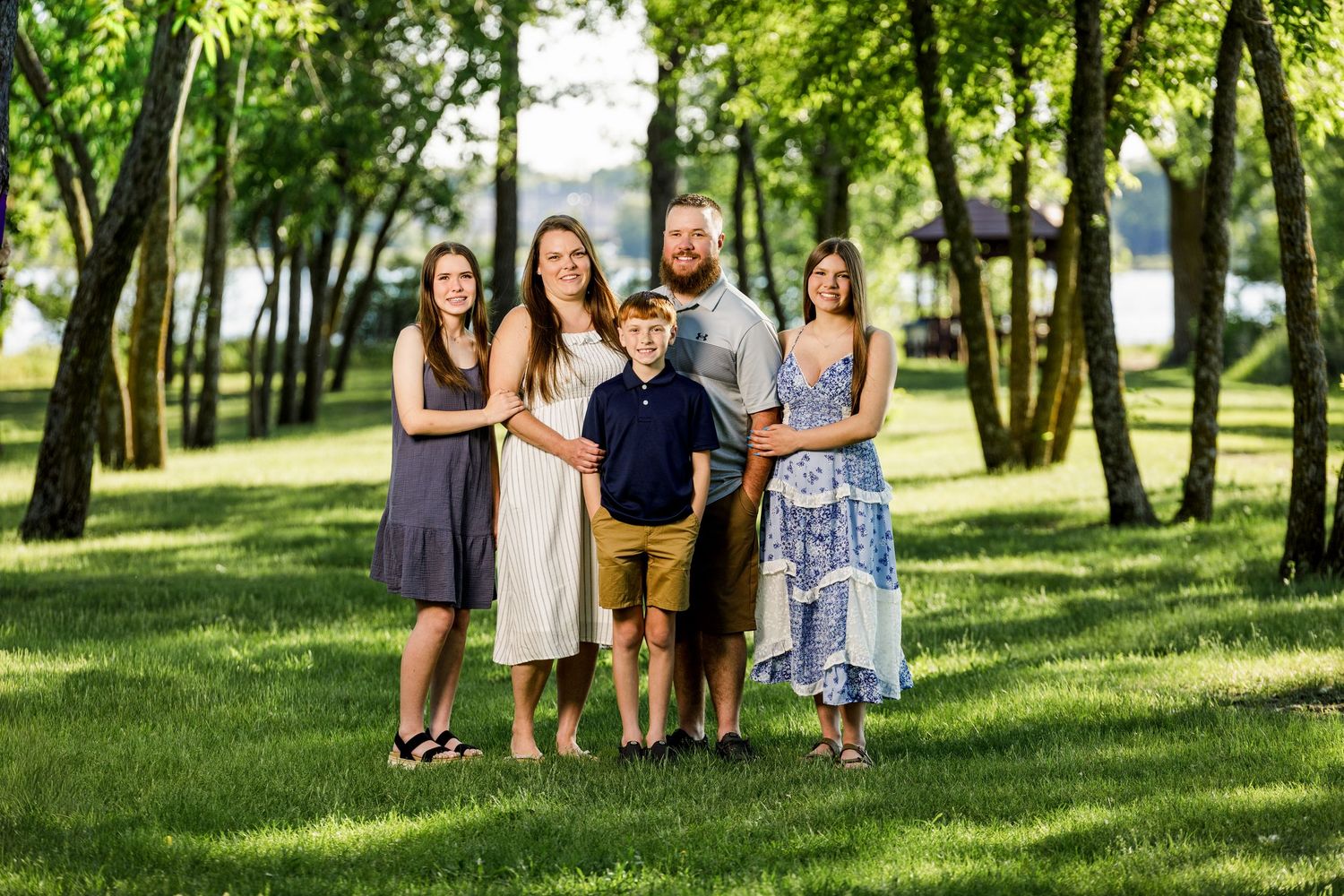 Family of five posing together in a lush green park with trees and a lake in the background.