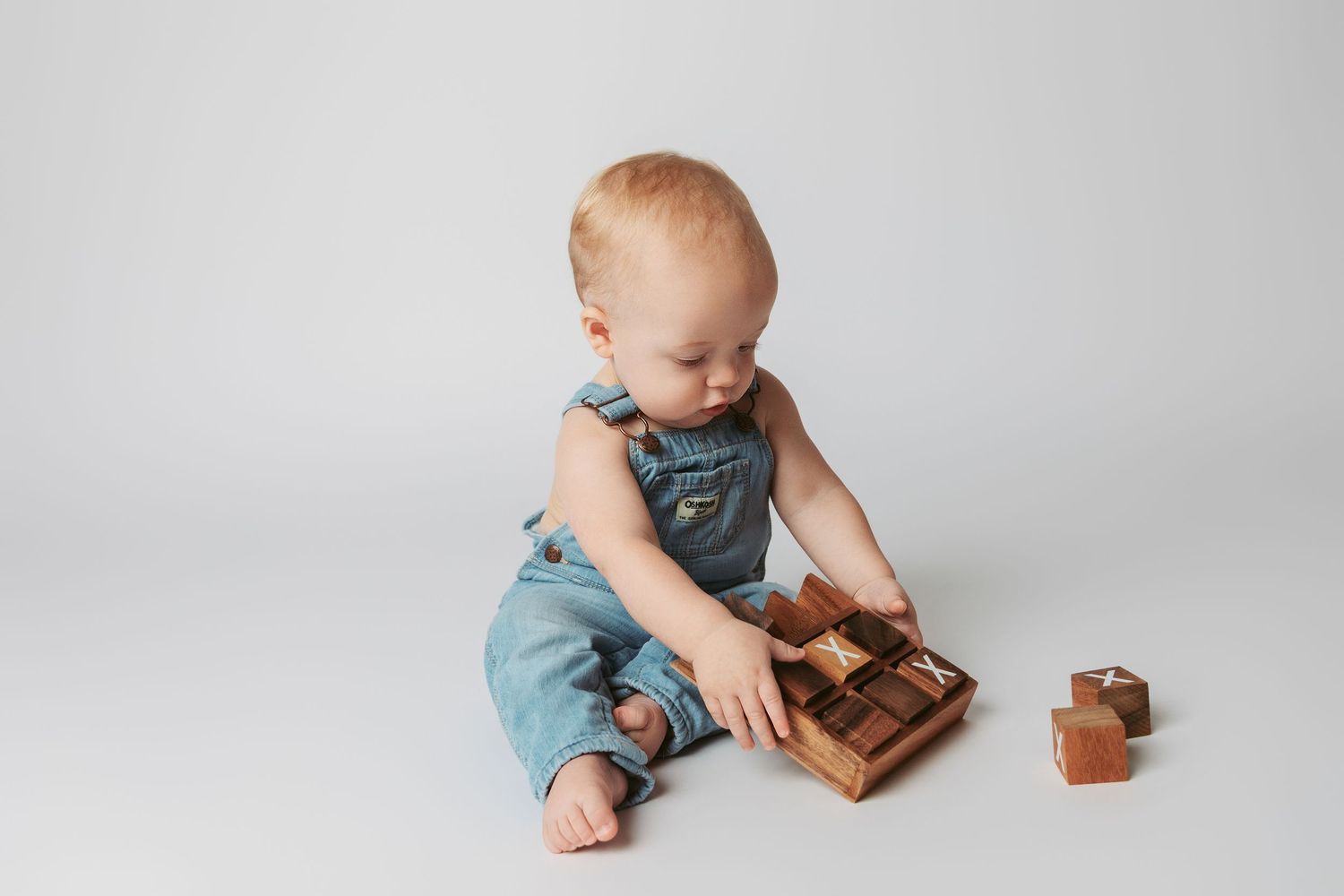 Small baby in blue overalls sits on white floor playing with wooden blocks and toy.