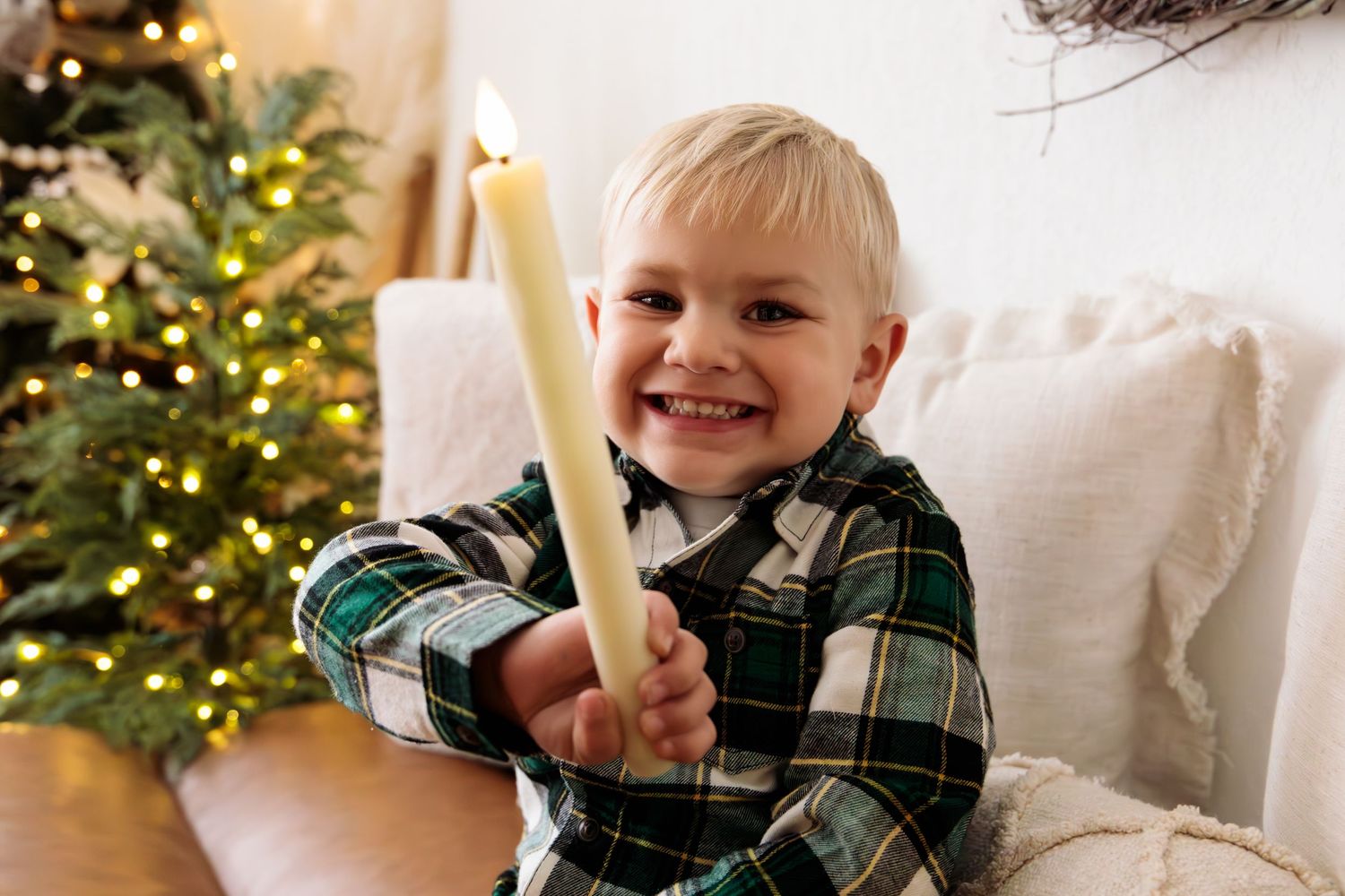 Child wearing plaid shirt smiles while sitting on white couch with Christmas tree in background.