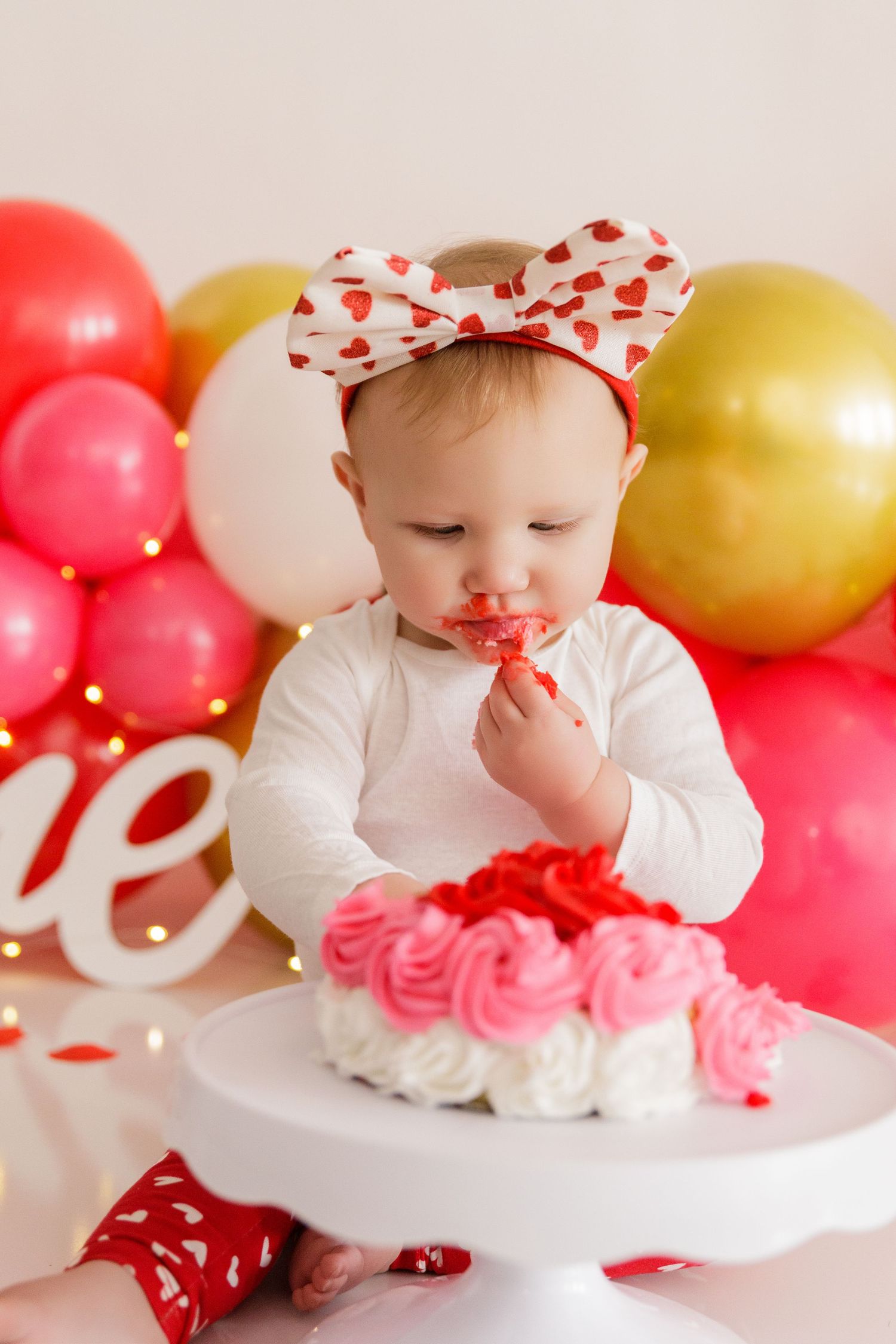 Valentine's themed cake smash setup with white cake stand and pink and gold balloon decorations.