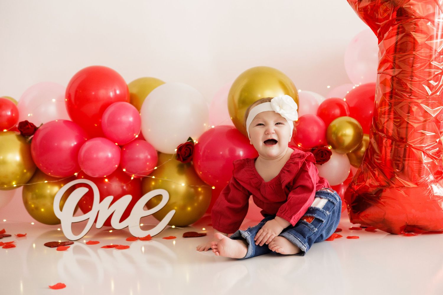 Baby in casual outfit lying on floor with red and gold balloon arch and 'one' sign decoration.
