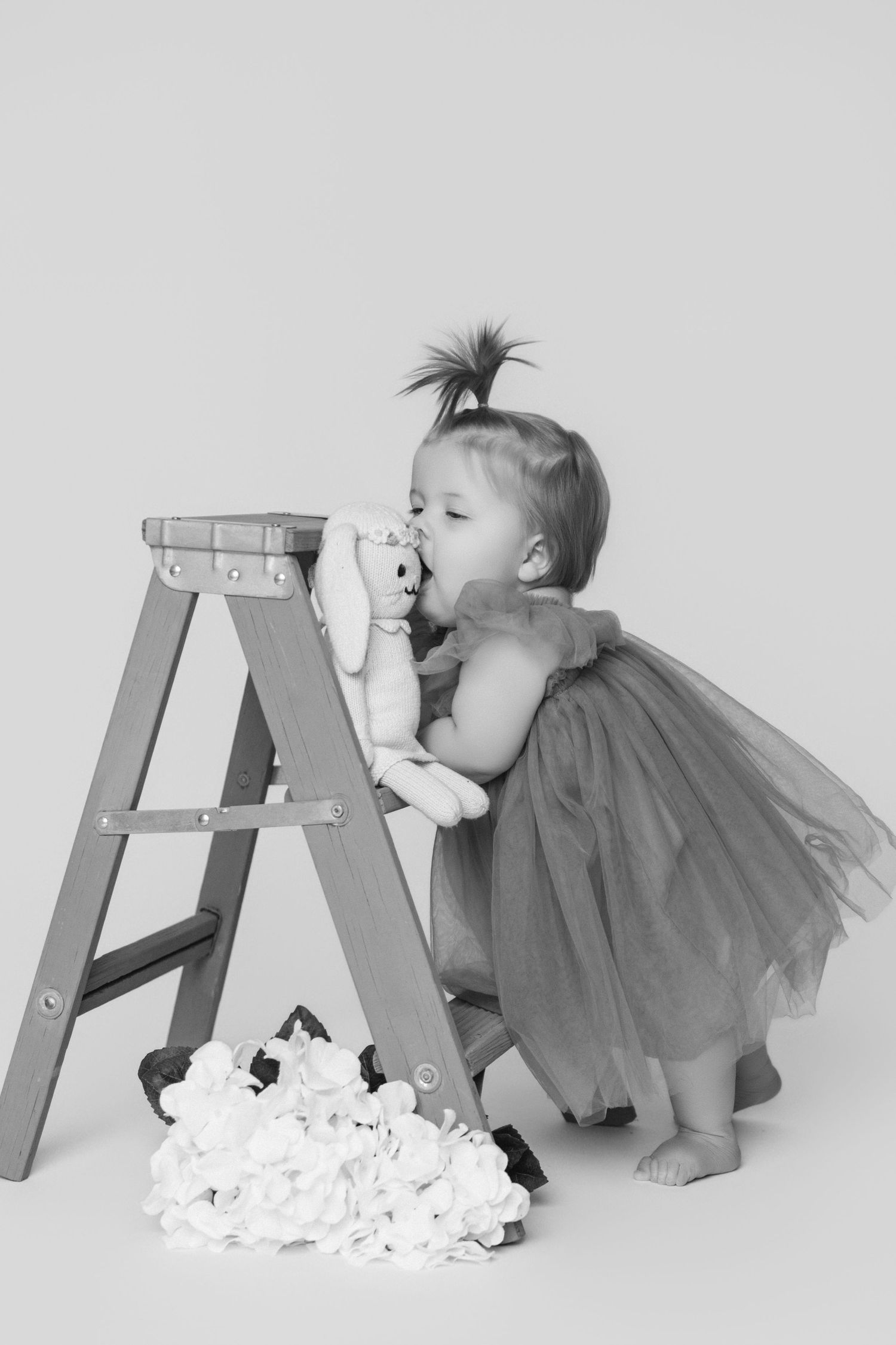 Black and white photo of baby in tulle dress reaching up to wooden ladder.