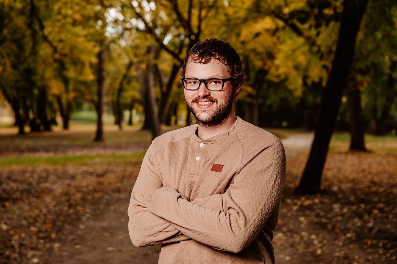 Person in glasses and beige shirt poses in autumn park setting with golden foliage in background.