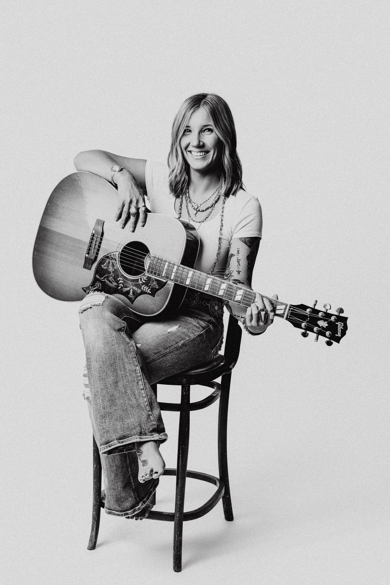 Black and white photo of a smiling woman sitting on a stool playing an acoustic guitar, wearing jeans and a white t-shirt.