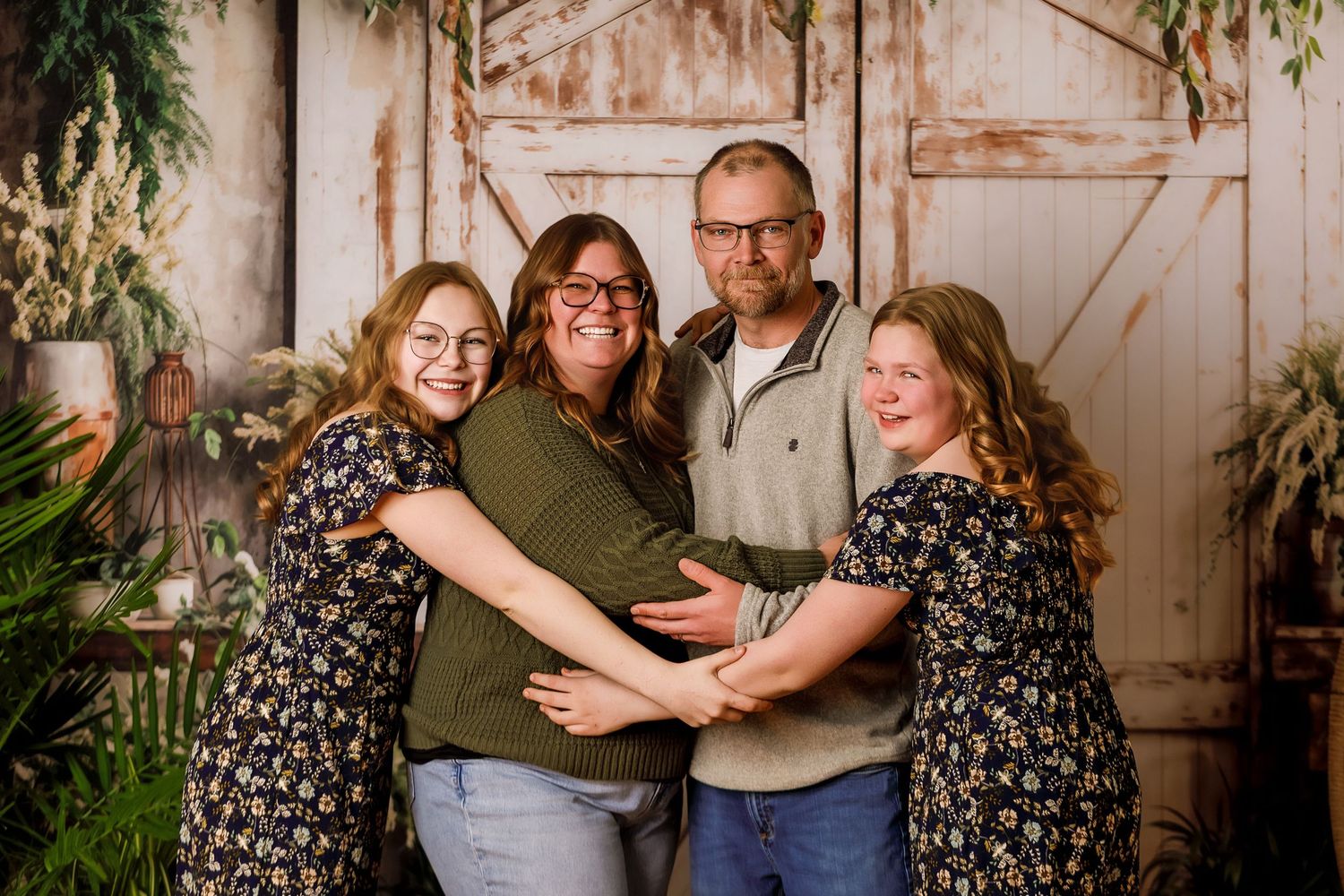 Family of four smiling and hugging in front of rustic barn doors with greenery backdrop in a professional photo session.