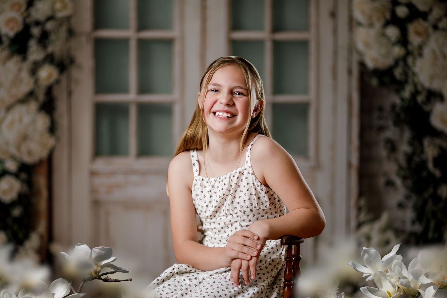 Smiling girl in floral dress sits before vintage door with white roses in elegant portrait photography.