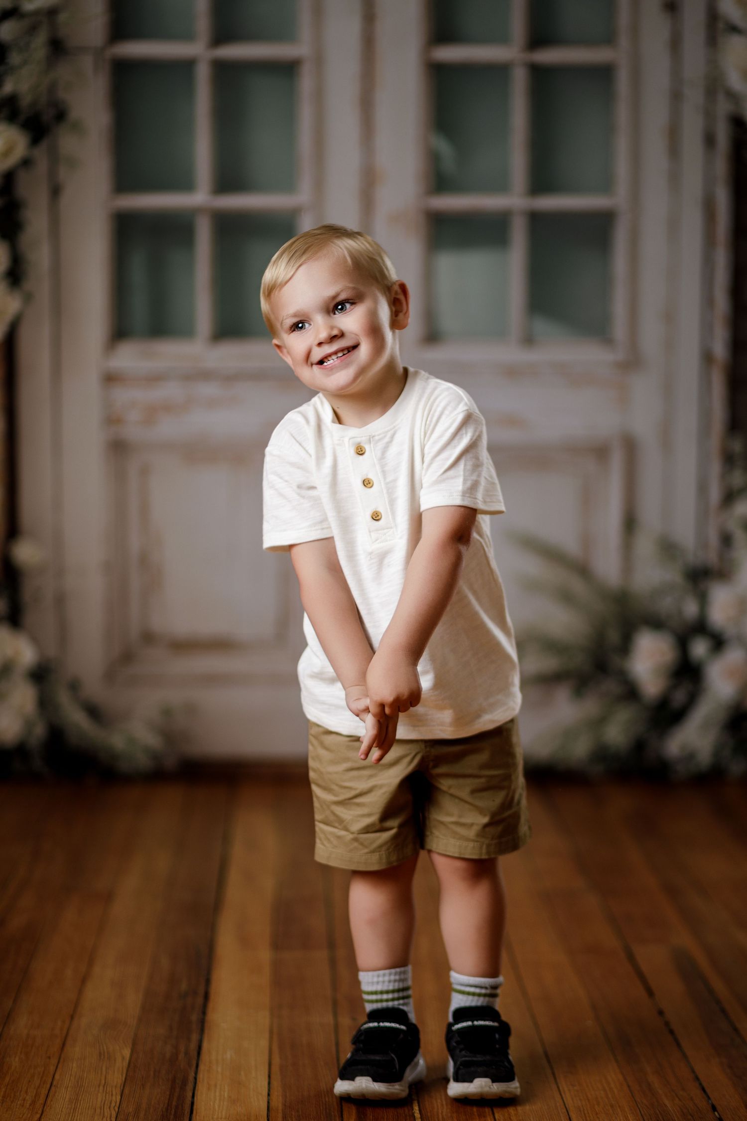Smiling young blonde boy in white shirt and khaki shorts standing on hardwood floor near vintage door.