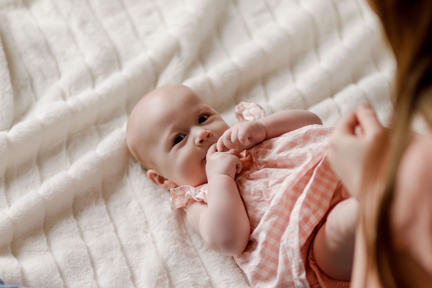Adorable baby in pink gingham outfit lying on white blanket with pacifier.