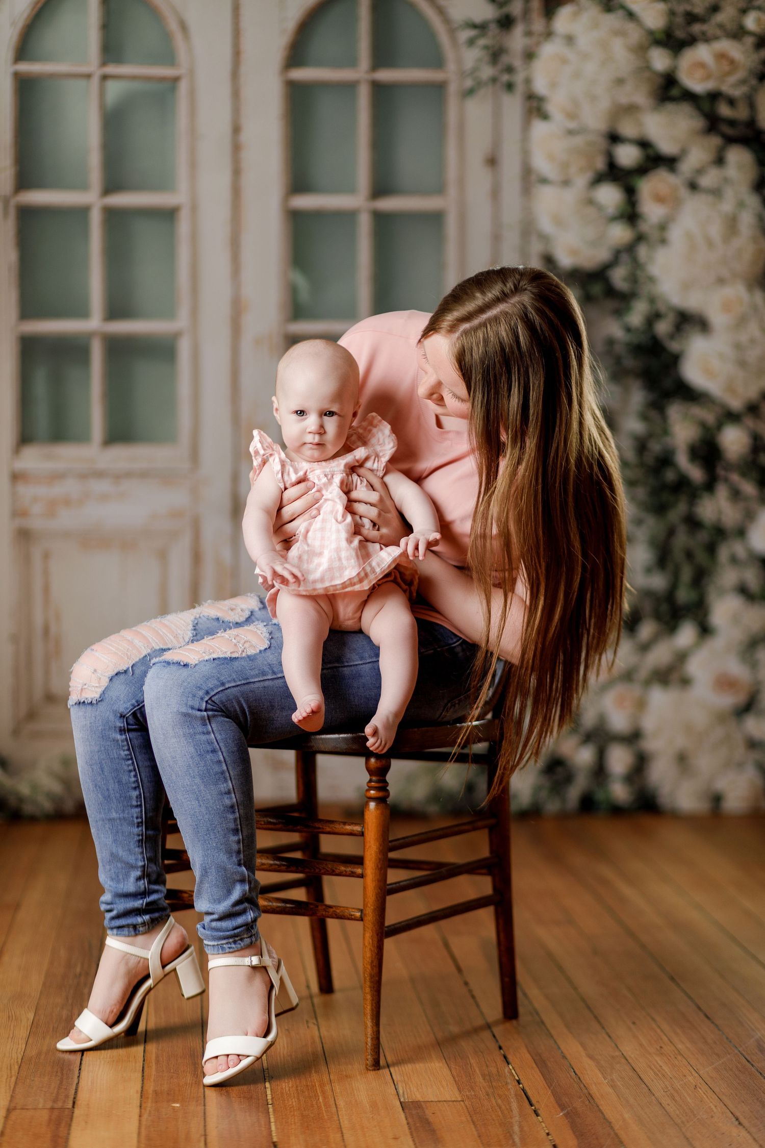 Mother in pink top and jeans sits on chair holding baby girl in pink dress in floral studio.