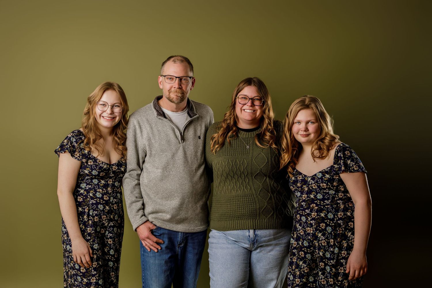 Family of four smiling together against an olive green background in a professional portrait studio photo.