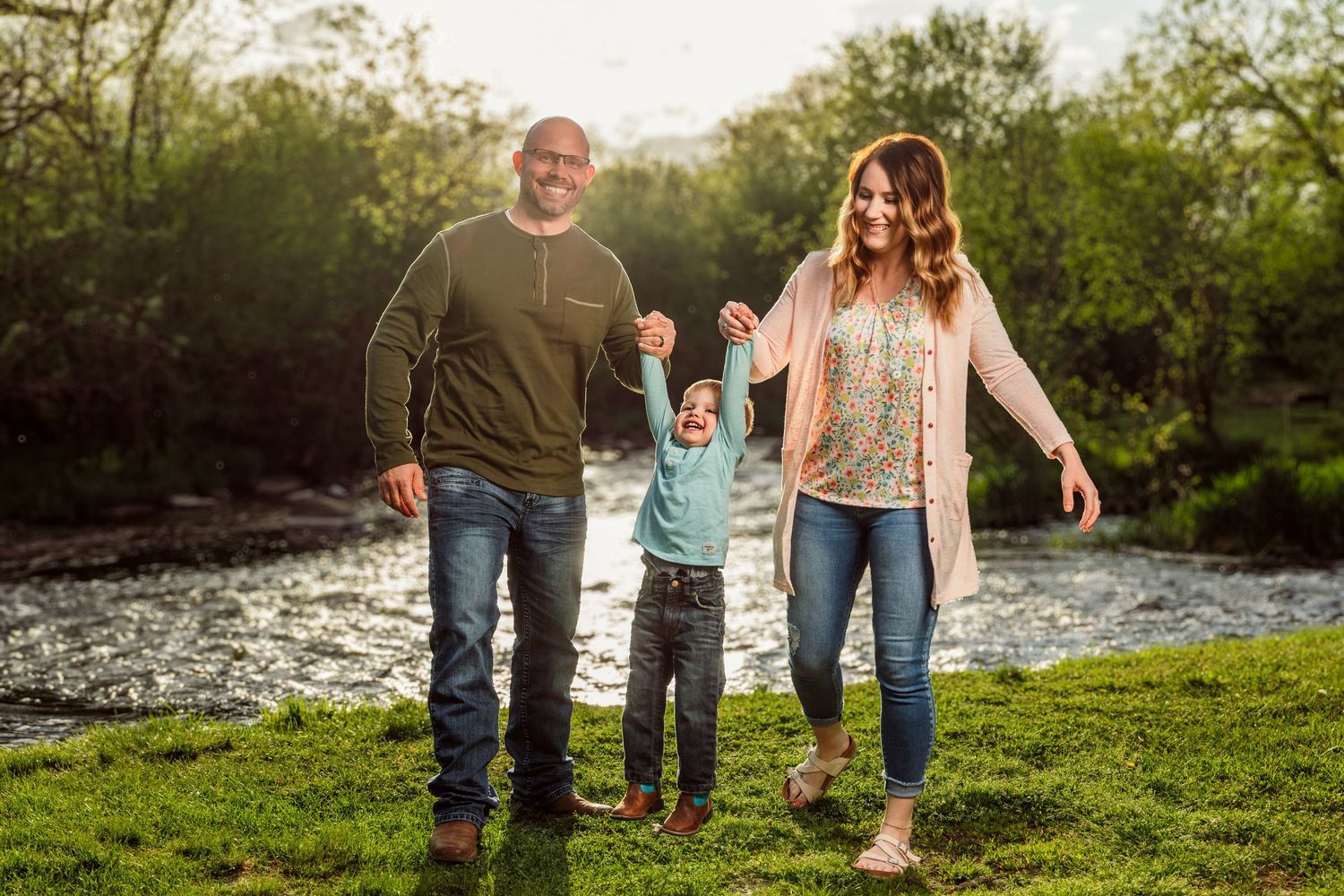 Family of three holding hands and walking by a sunlit river on green grass surrounded by trees.