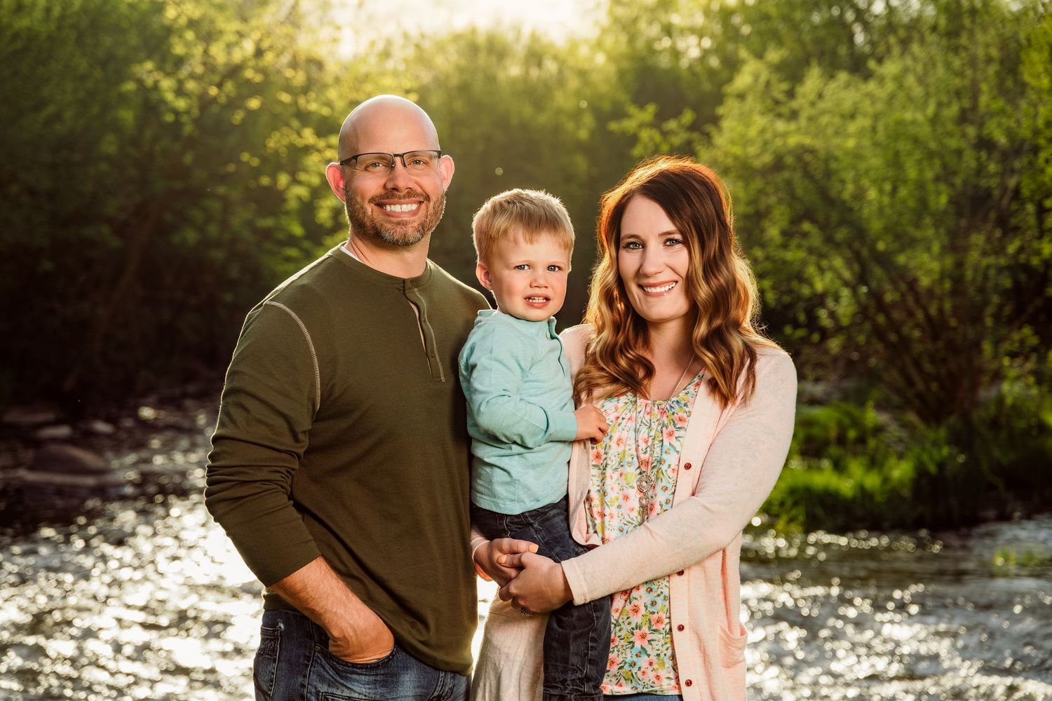 Happy family portrait with parents and toddler son smiling outdoors near a river with lush green trees.