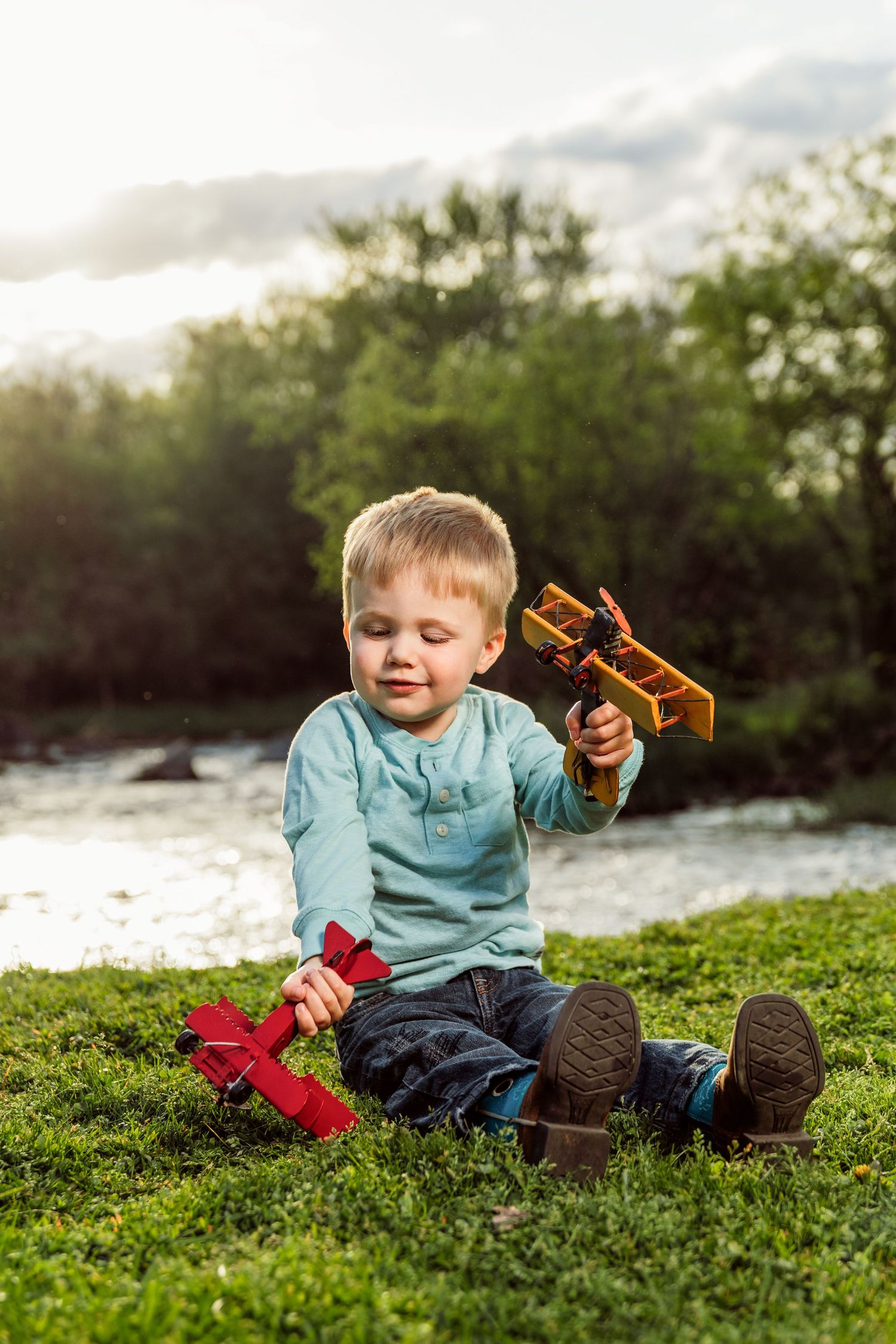 Young boy sitting on grass playing with wooden and red toy airplanes near a river on a sunny day.