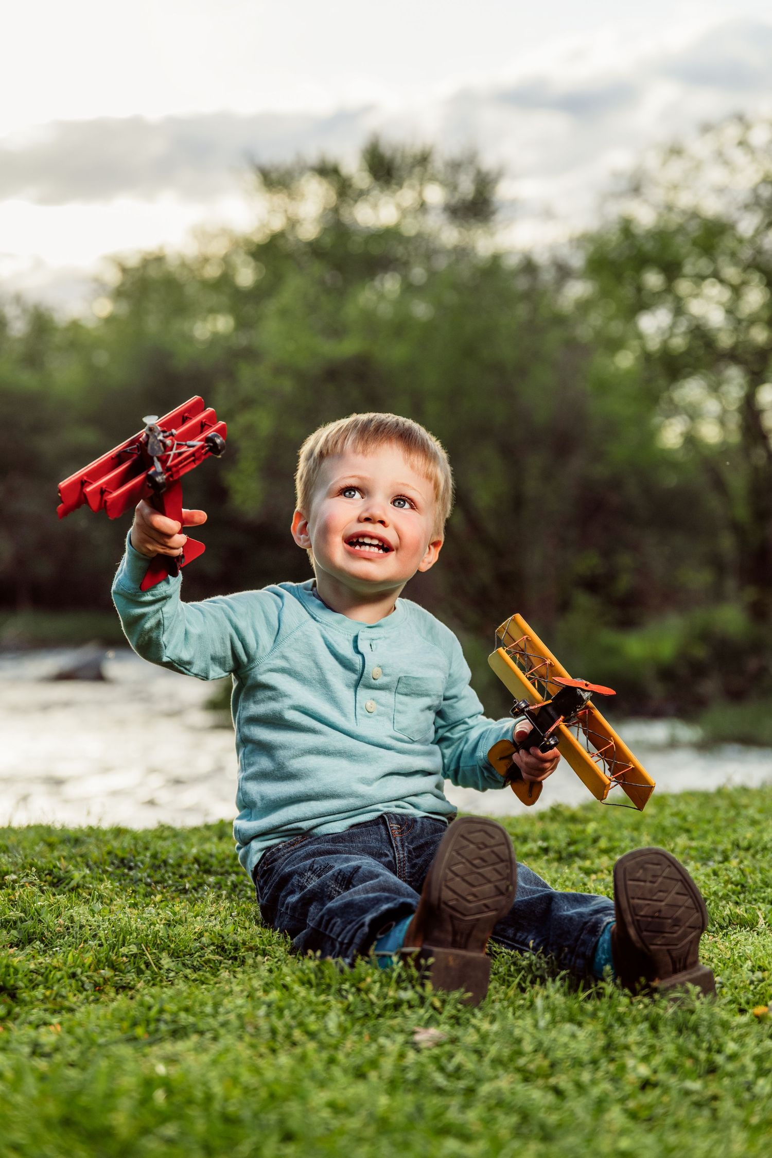 Young boy sitting on grass holding two toy airplanes outdoors, smiling joyfully with trees and river in background.