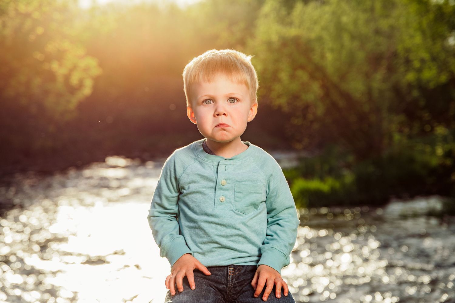 A young blond toddler boy in a teal shirt sits outdoors near a sunlit river surrounded by green trees.