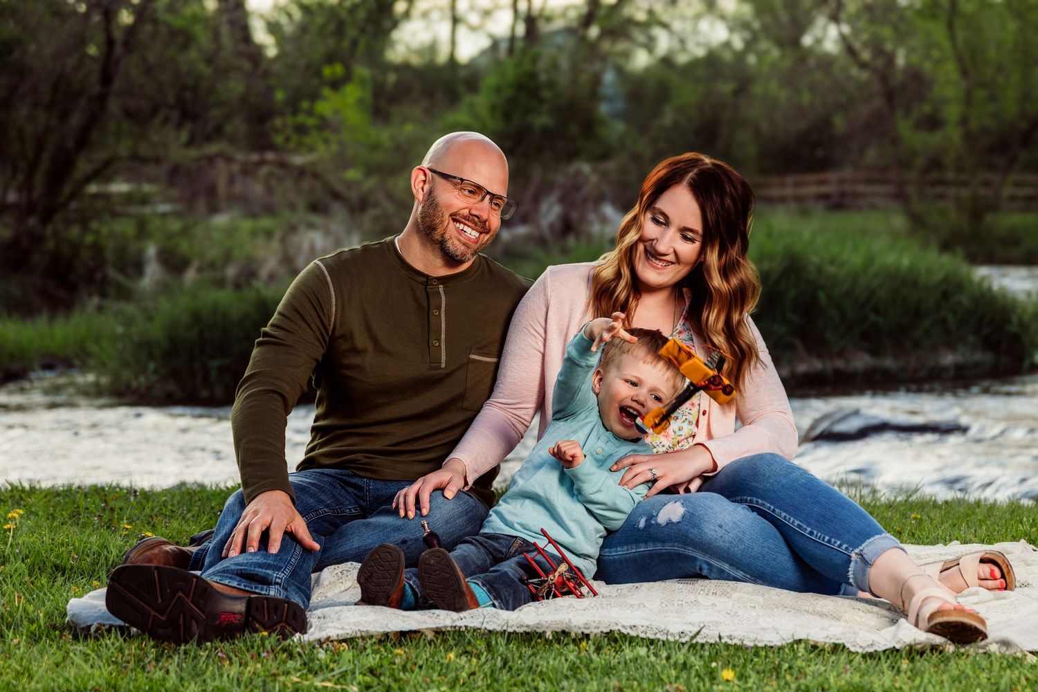 Happy family of three sitting on a blanket outdoors near a river, smiling parents with their young toddler child.