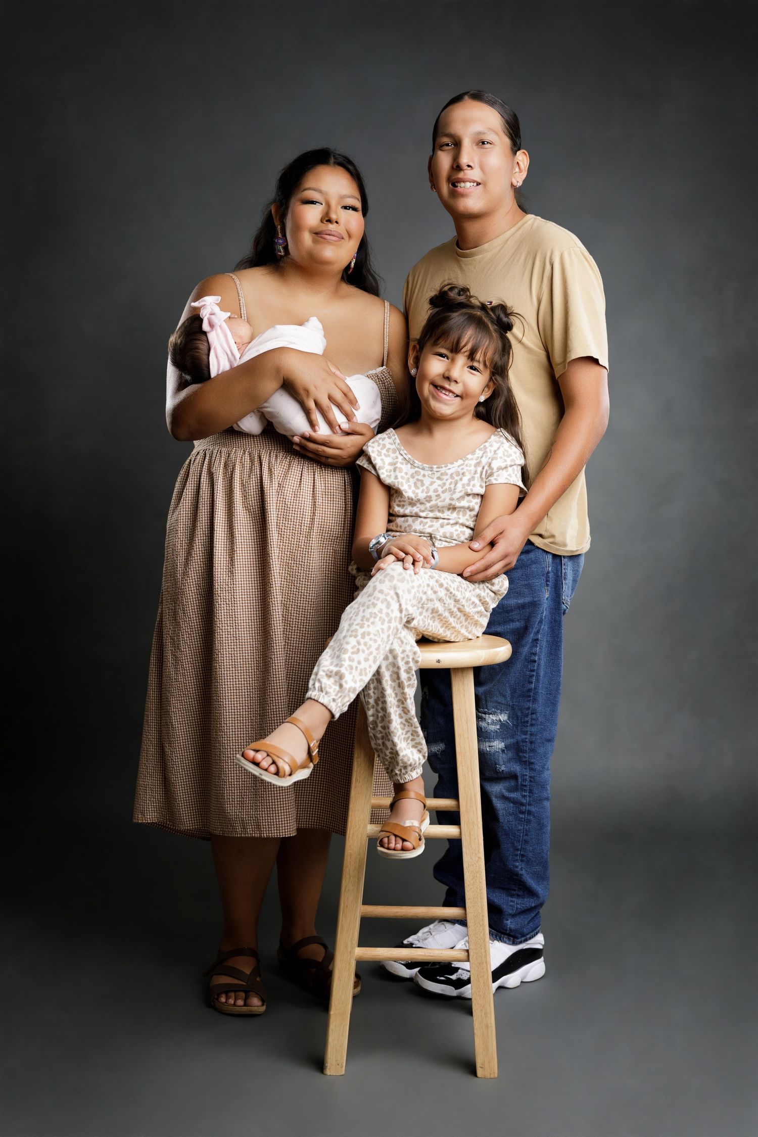Family portrait of parents with young daughter seated on stool and newborn baby in professional studio setting.