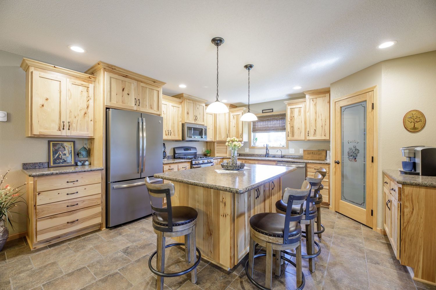 Modern kitchen with light wood cabinets, granite island, bar stools, stainless steel appliances, and pendant lighting.