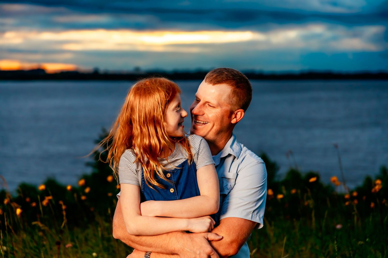 Couple laughs together in a sweet moment by the water during golden hour with dramatic clouds overhead.