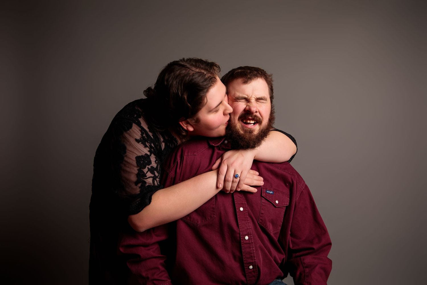 A couple shares a tender embrace during an engagement photo session against a neutral backdrop.