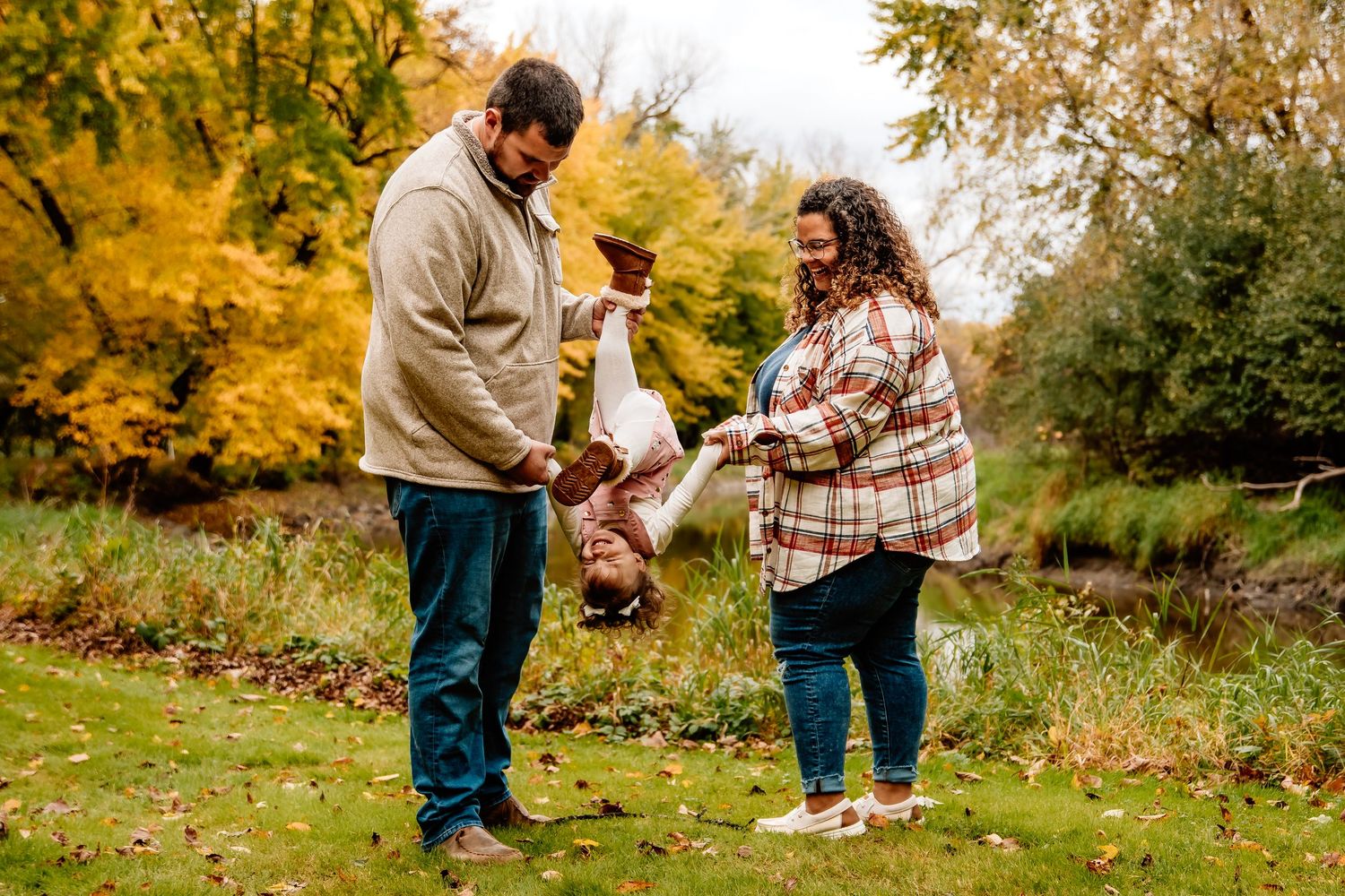 A couple swings a small child by their arms while surrounded by autumn foliage in a park setting.