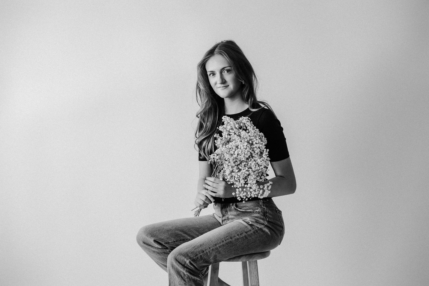 Young woman in floral top and jeans sitting on stool, black and white studio portrait photography.