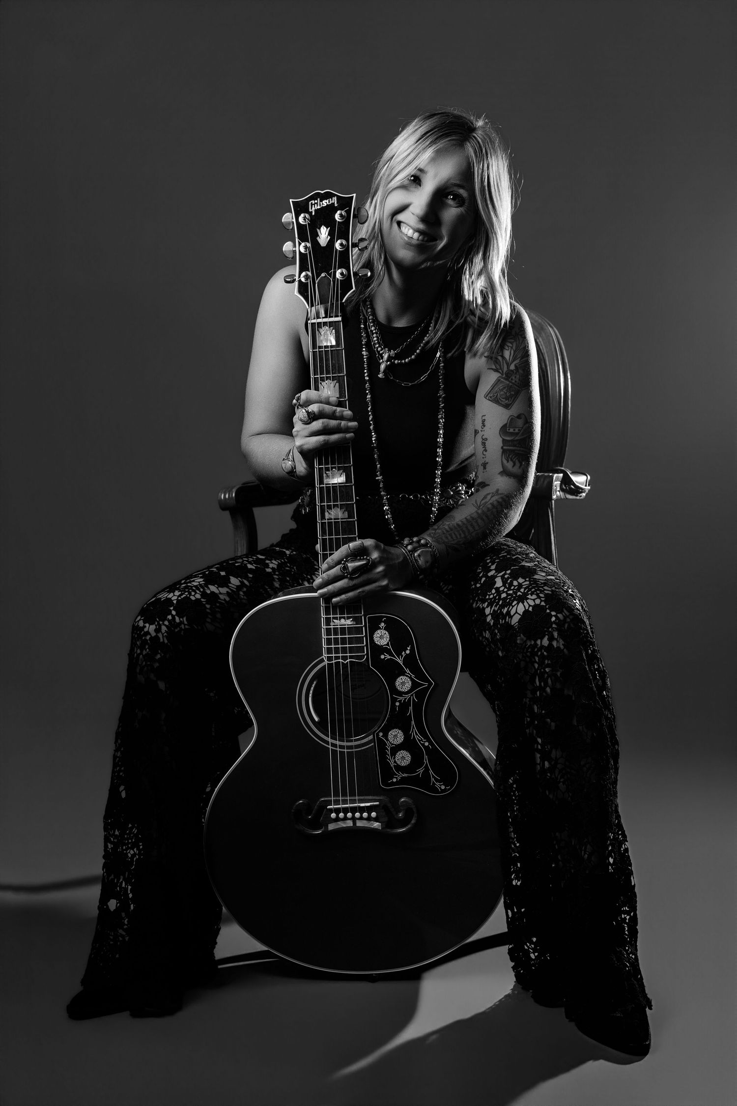Black and white portrait of tattooed female guitarist holding Gibson acoustic guitar while seated.