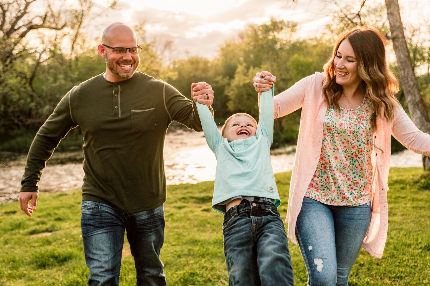 Happy family with parents swinging young child outdoors in green park at golden hour sunset.