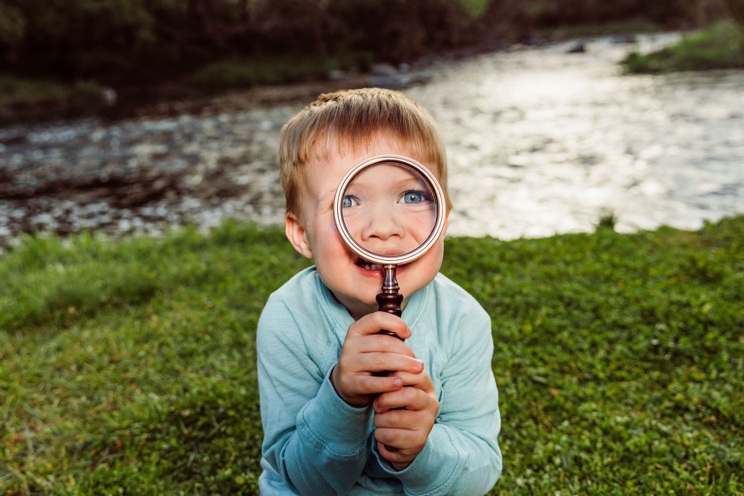 Curious young boy holding a magnifying glass up to his face near a river, enlarging his eye outdoors.