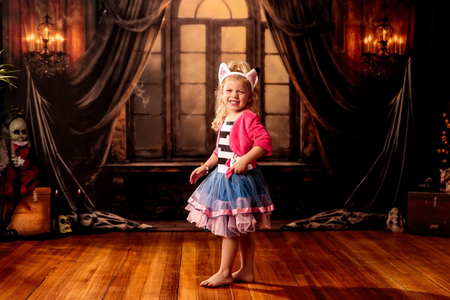 Young girl in pink cardigan and layered tutu dress poses barefoot on wood floor before ornate vintage backdrop.