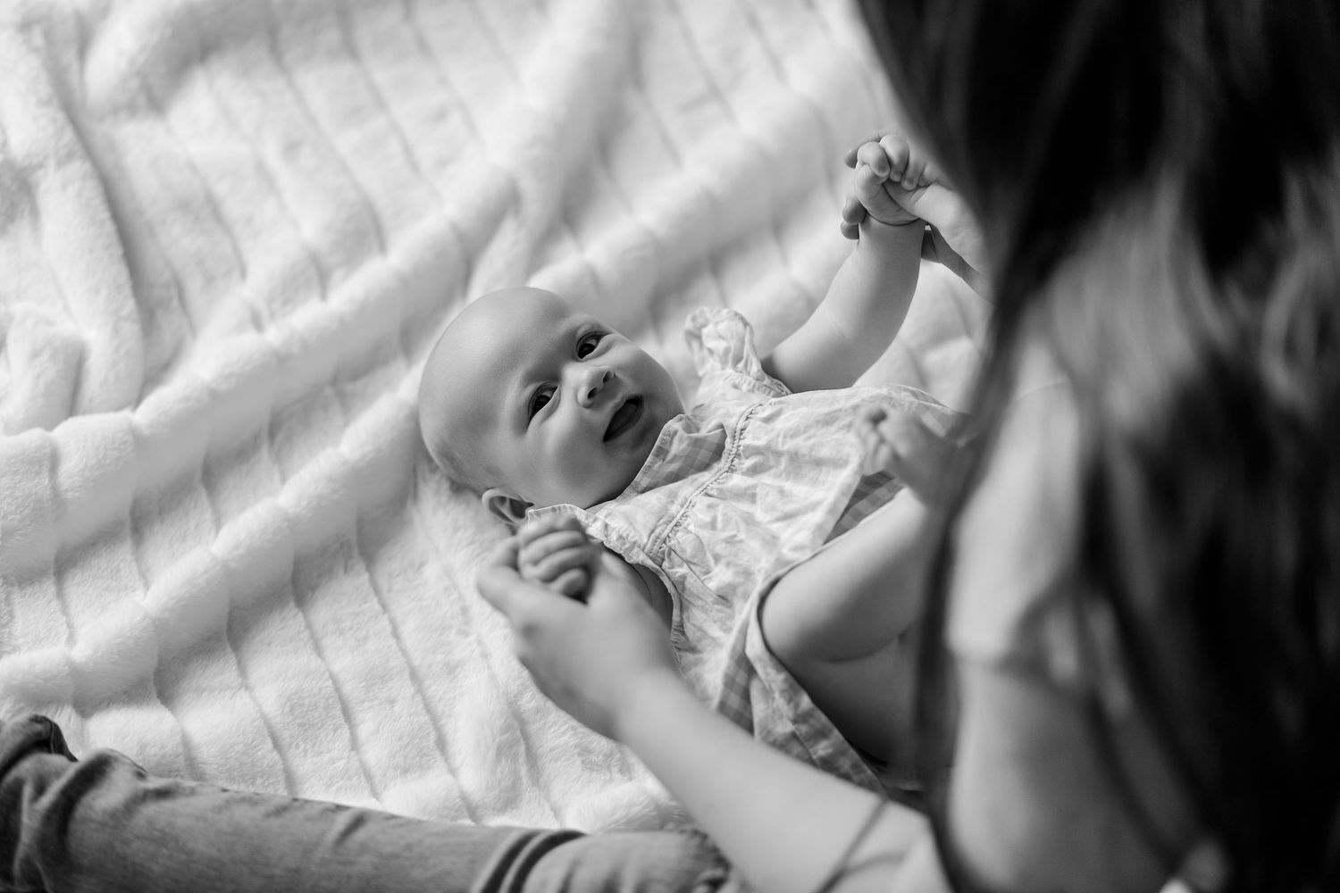 Black and white photo of mother interacting with smiling baby lying on white blanket.