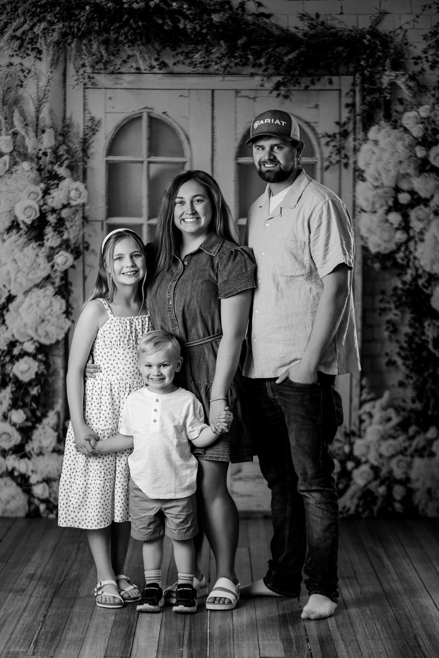 Black and white family portrait of parents and two young children posing in front of floral backdrop and vintage doors.