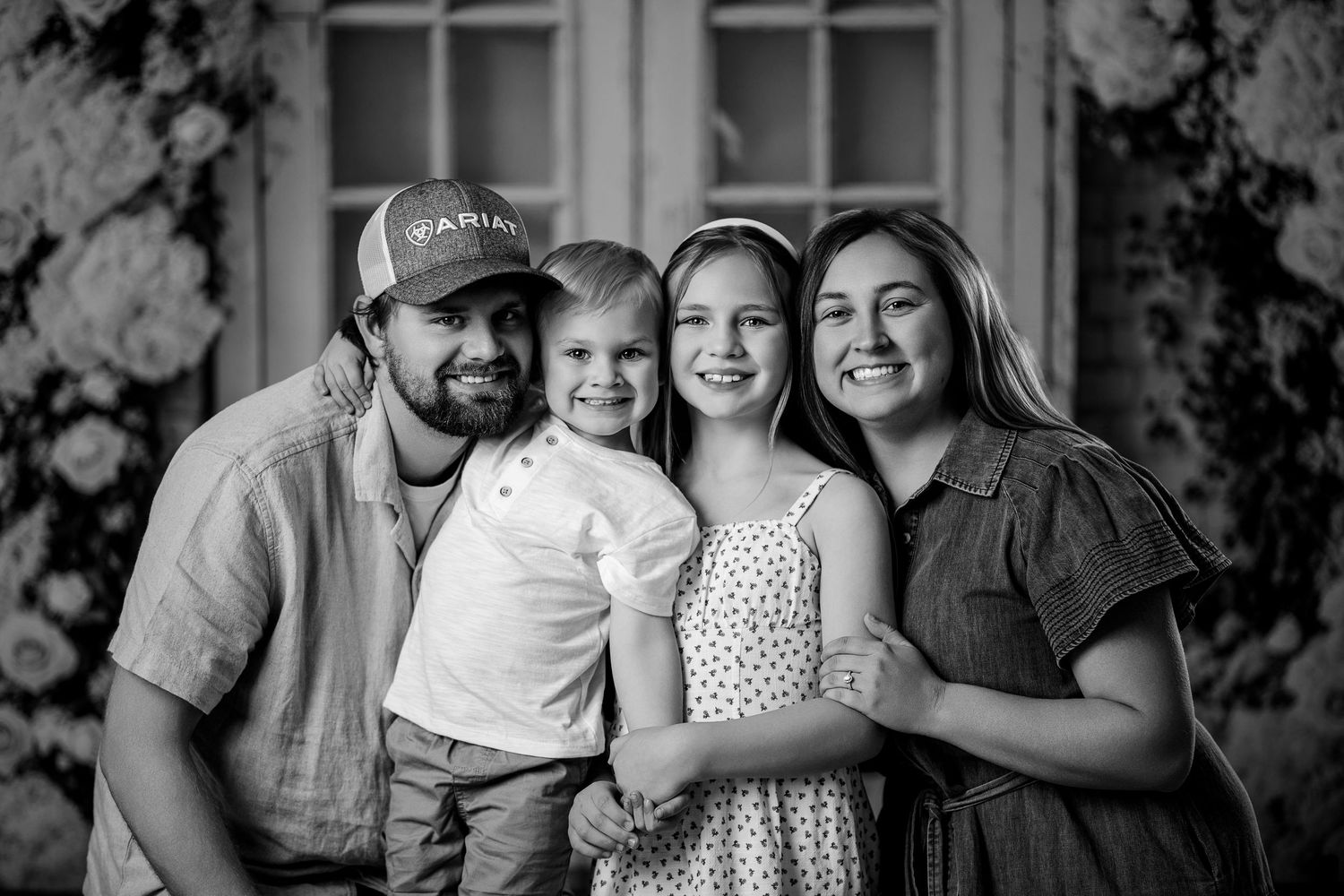 Black and white family portrait of parents and two young children smiling together in front of floral backdrop.