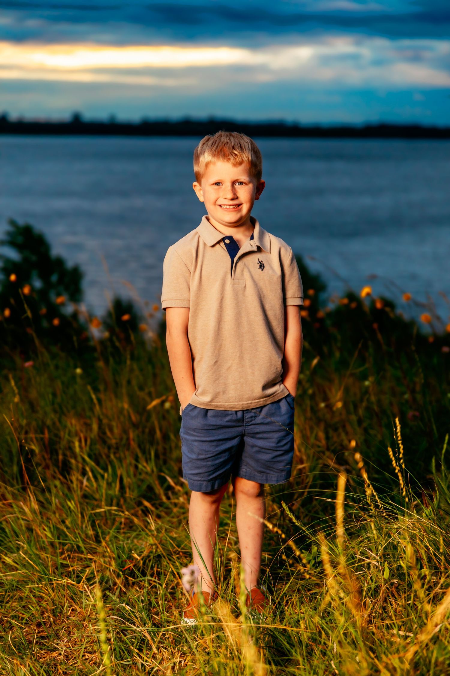 Young child standing in tall grass by a lake at sunset wearing casual summer clothing.
