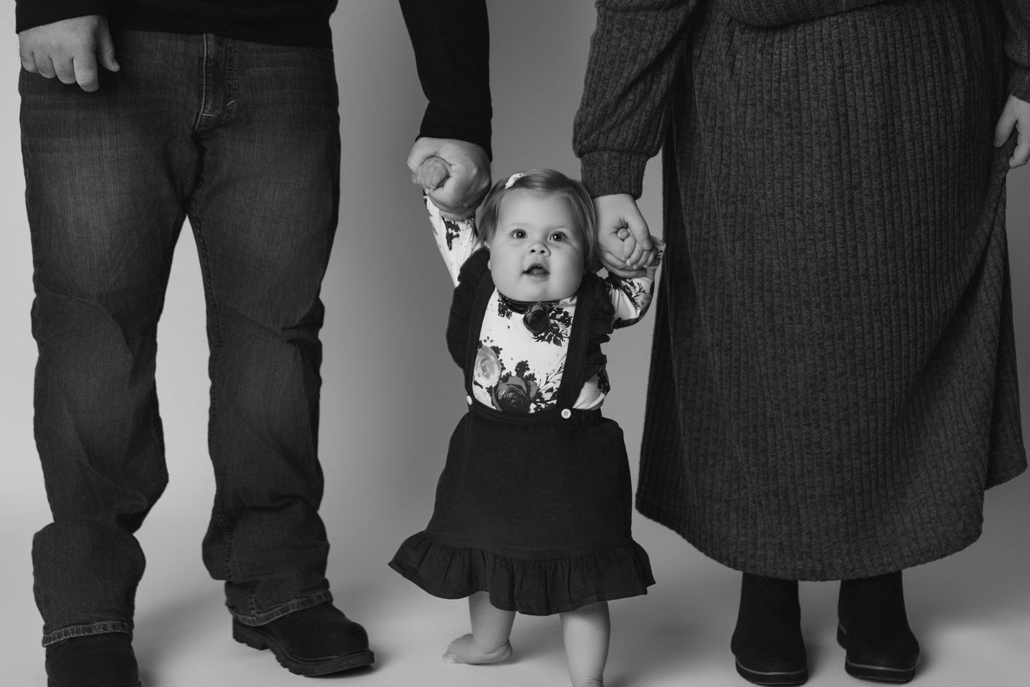 Black and white photo of a baby walking while holding hands with adults.