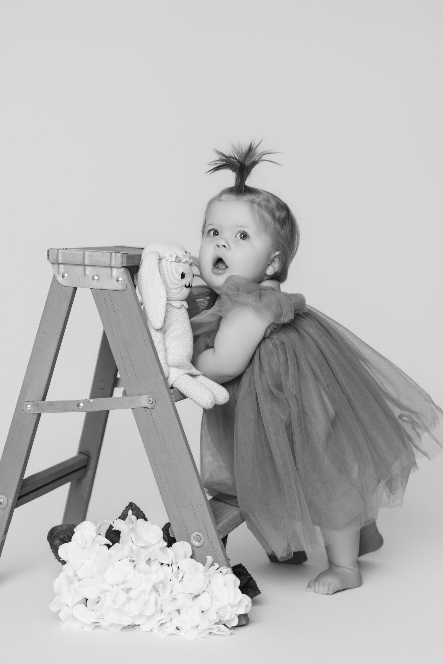 A toddler in a tulle dress curiously peers at a stepladder with flowers scattered below.