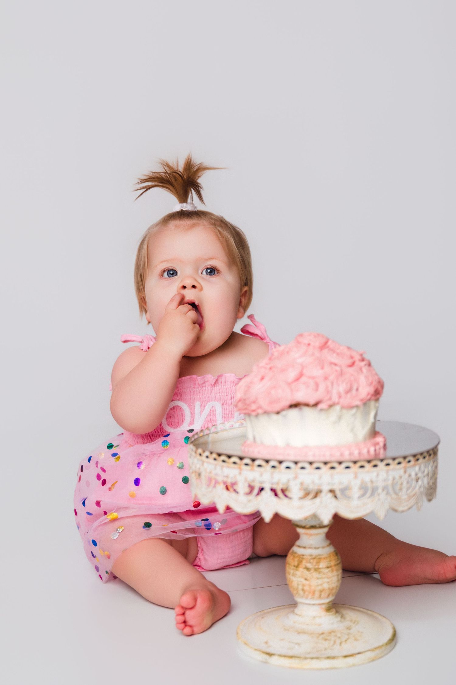 Baby in pink polka dot outfit reaching for cake on decorative white cake stand.