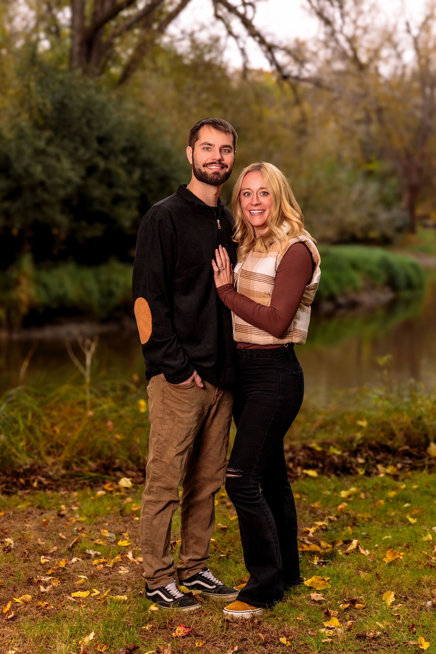 Couple poses playfully in a fall setting with autumn leaves on the ground near a pond.
