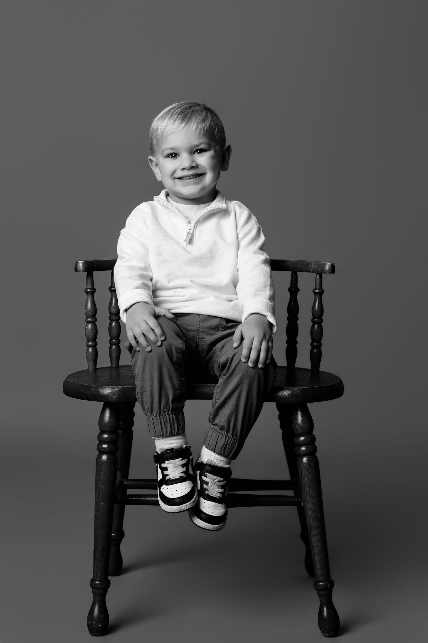 A small child sits playfully on a dark wooden bench against a gray background.