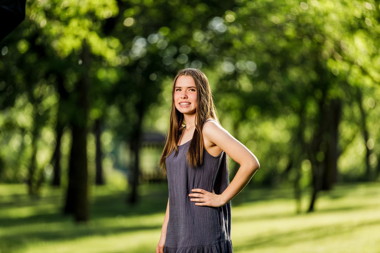 Young teenage girl in a gray dress posing with hand on hip in a sunlit green park.