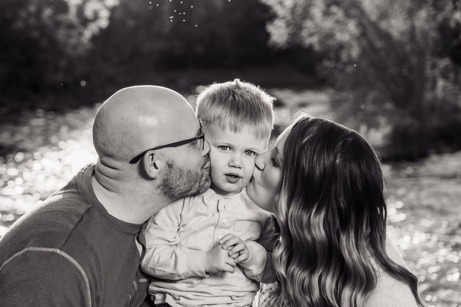 Black and white photo of parents kissing their toddler son on the cheeks outdoors in nature.