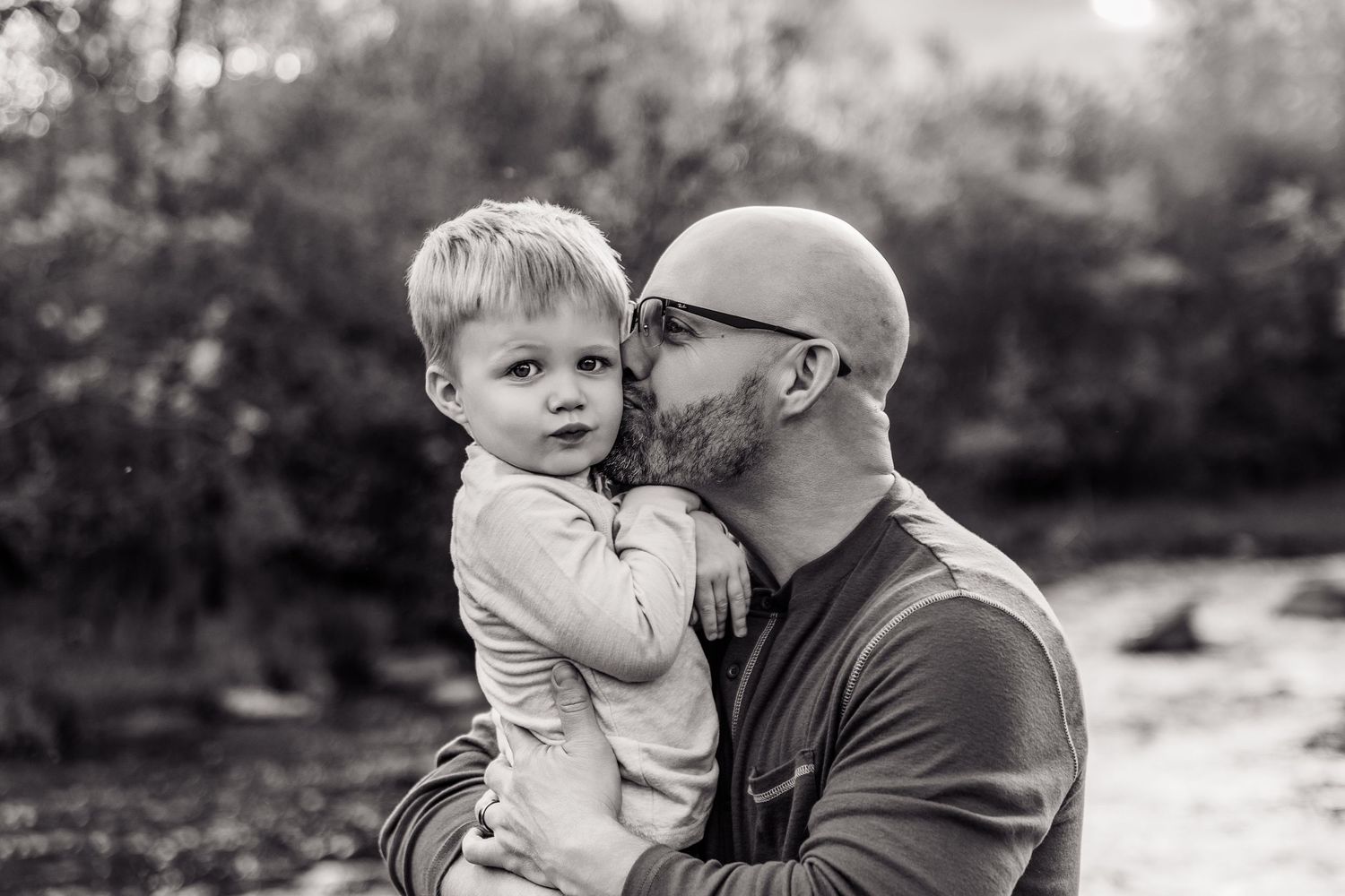Black and white photo of a bald father kissing his young toddler son outdoors near a river.