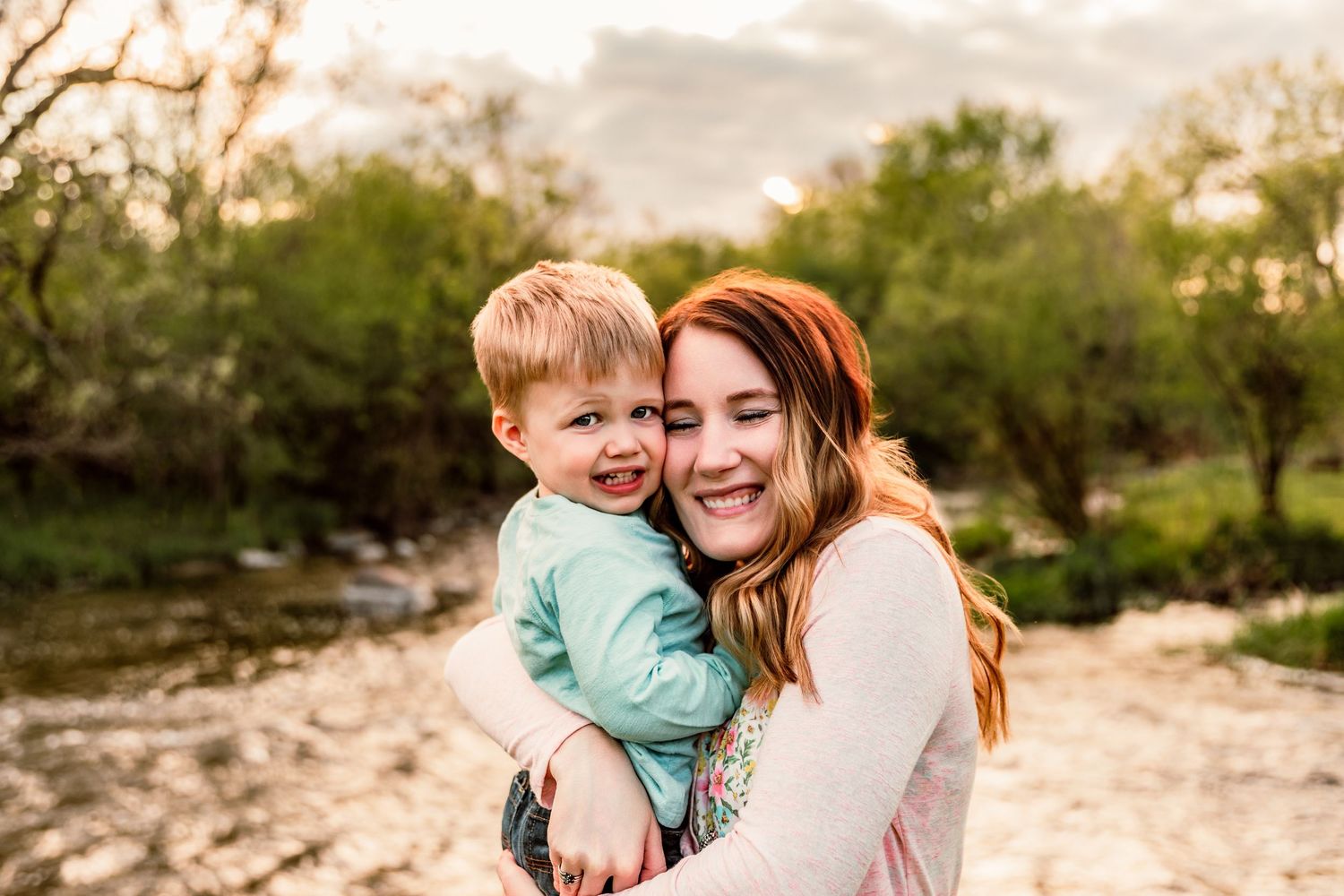 A smiling redhead woman hugs a happy toddler boy outdoors near a creek with lush green trees at sunset.